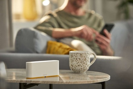  Na stolku stojí TaHoma Somfy – centrální jednotka smart home. V pozadí muž používá smartphone.